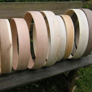 Puede incluir: Una serie de nueve aros de madera circulares dispuestos en fila, que varían en diámetro y patrones de veta de madera. Los aros están construidos con madera natural de color claro, con un aro que presenta un tono de madera grisáceo-marrón más oscuro.