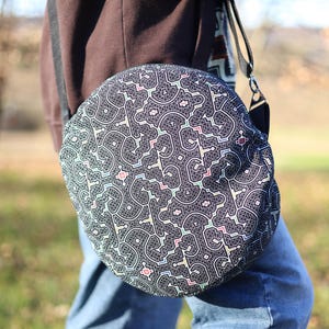 Könnte beinhalten: Eine runde, schwarze Umhängetasche mit einem sich wiederholenden geometrischen Muster in Weiß, Rosa und Blau. Die Tasche hat einen schwarzen Riemen und wird über der Schulter getragen. Die Person trägt eine blaue Jeans und eine braune Jacke.