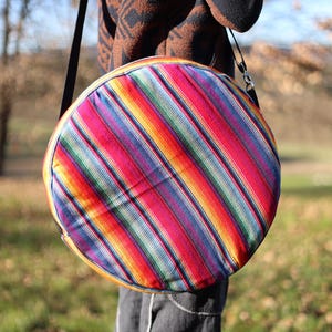 Könnte beinhalten: Eine runde, farbenfrohe Umhängetasche mit einem gestreiften Muster in Rot-, Orange-, Gelb-, Grün- und Blautönen. Die Tasche hat einen schwarzen Riemen und eine gelbe Einfassung. Die Tasche wird von einer Person getragen, die einen braun-schwarz gemusterten Pullover und dunkelgraue Hosen trägt.