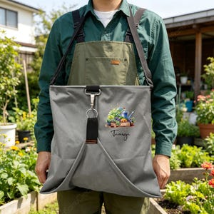 May include: A gray canvas apron with black adjustable straps and a central metal clip. The front features a printed illustration of a wooden crate filled with garden plants and a blue watering can, with the name Tamsyn written in black script.