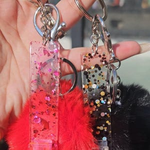Può includere: Due portachiavi con ciondoli in resina trasparente pieni di glitter. Un ciondolo è rosa e rosso con un pon pon rosso soffice. L'altro ciondolo è nero con glitter dorati e bianchi e un pon pon nero soffice. Entrambi i ciondoli hanno un clip in plastica trasparente.