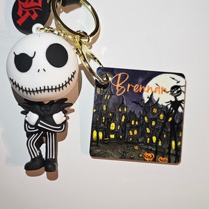 Puede incluir: Un llavero de Jack Skellington en blanco y negro con una cadena dorada y una correa negra. El llavero también incluye un pequeño amuleto rojo con un carácter kanji negro y un amuleto cuadrado con una imagen en blanco y negro de Jack Skellington frente a un pueblo espeluznante con el nombre "Brennan" escrito en naranja.