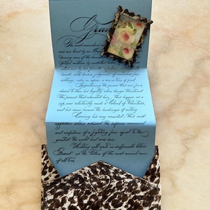 Könnte beinhalten: Eine hellblaue Karte mit handschriftlichem Text und einem kleinen gerahmten Blumendruck. Die Karte ist gefaltet und zeigt mehr Text im Inneren. Der Umschlag besteht aus einem braunen und weißen Tierdruckstoff.
