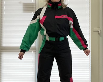 May include: A vintage black and color block ski suit. The suit features green, white, and pink accents on the sleeves and legs. It has a high collar, a belt, and a zipper closure. The suit is designed for cold weather activities.