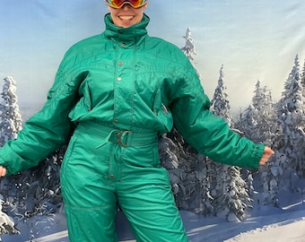 May include: Emerald green vintage ski suit with a high collar, button details, and a belted waist. The one-piece suit has long sleeves and pants. The person is wearing ski goggles with a reflective lens.