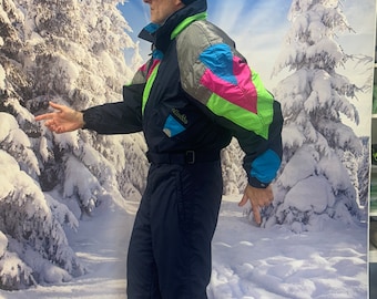 May include: A blue and black ski suit with a colorful geometric pattern on the jacket. The jacket has a hood and a high collar. The suit is worn by a person wearing a black ski hat with the word "ellesse" on it and ski goggles.