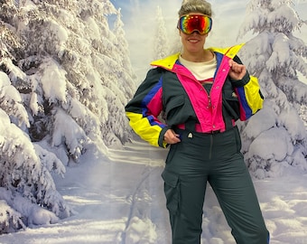 May include: A woman wearing a bright pink, blue, and yellow ski suit with a black base layer. She is wearing rainbow ski goggles and standing in a snowy forest setting.