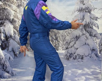 May include: A blue one-piece ski suit featuring purple and yellow color-blocked accents on the shoulders and sleeves. The suit includes a high collar, a cinched waist with a belt, and long sleeves. A red beanie and ski goggles are worn.