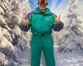 May include: A teal and purple ski suit with a black belt and a black hat. The ski suit has the brand name "FILA" on the chest. The person is wearing orange and red ski goggles.