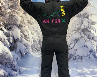 May include: Black one-piece ski suit with the text "K-WAY ART FUN" in green and pink. The suit has a black belt and a blue beanie with a silver stripe. The background features a snowy winter scene.