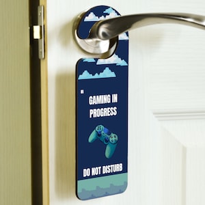 May include: A blue door hanger with a pixelated design of clouds and a video game controller. The text "Gaming in Progress" and "Do Not Disturb" is printed on the hanger.