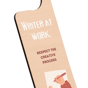 Può includere: Un appendiporta in legno con il testo "Writer at Work. Respect the Creative Process" e un'illustrazione di mani che scrivono su un foglio di carta.