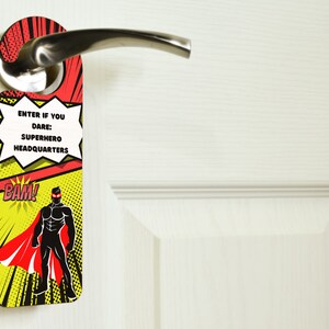 Op de afbeelding: Een rode en gele deurhanger met een superheldensilhouet en de tekst "Kom binnen als je durft: Superheldenhoofdkwartier" en "BAM!"