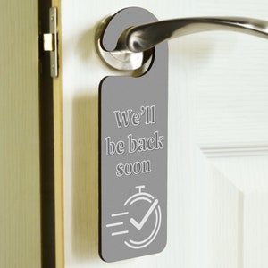 Può includere: Appendino per porta grigio con il testo "We'll be back soon" e un segno di spunta bianco all'interno di un cerchio con tre linee sopra.