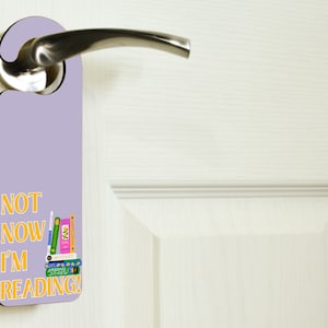 Könnte beinhalten: Ein lilafarbener Türhänger mit dem Text "NOT NOW I'M READING!" in gelben Buchstaben. Neben dem Text ist ein Stapel bunter Bücher abgebildet.