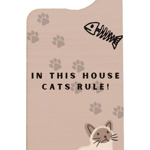 May include: A light brown wooden door hanger with a white cat illustration and the text "In this house cats rule!"  The hanger has a hook at the top and paw prints scattered around the cat.