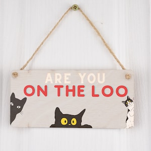 Può includere: Un cartello di legno con il testo "Are you on the loo" in lettere rosse e blu. Il cartello ha tre gatti neri che spuntano da dietro.