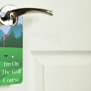 May include: A green and blue door hanger with a golf course scene and the text "I'm On The Golf Course".
