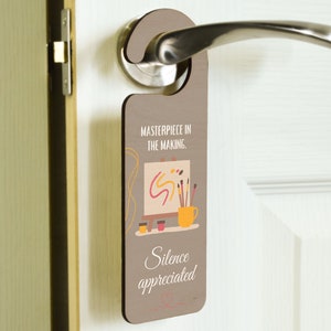 May include: Brown wooden door hanger with the text "Masterpiece in the making. Silence appreciated." and a cartoon illustration of an easel with paintbrushes and a cup of paint.