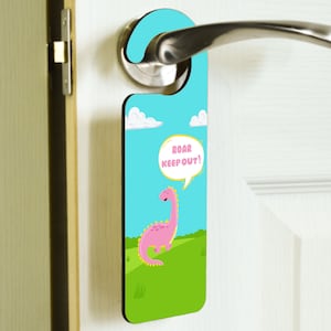 以下が含まれることがあります： 白い雲のある青い背景に、黄色い斑点のあるピンクの恐竜がいます。恐竜は「Roar Keep Out!」と書かれた吹き出しを持っています。恐竜は緑の草原に立っています。これはドアハンガーです。
