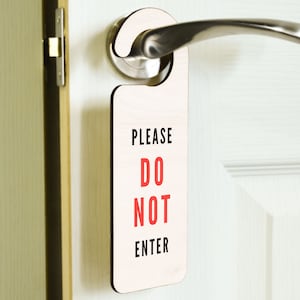 Puede incluir: Un colgador de puerta de madera blanco con un borde negro y el texto "PLEASE DO NOT ENTER" en letras rojas y negras.