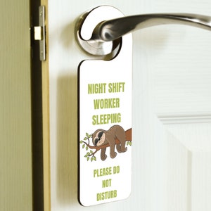 May include: A white door hanger with a cartoon sloth sleeping on a branch. The text on the hanger reads "Night Shift Worker Sleeping Please Do Not Disturb".