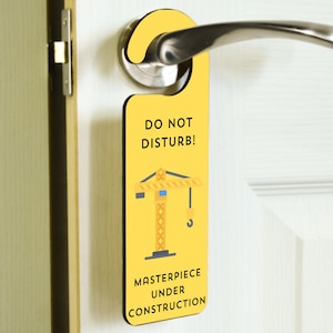 Puede incluir: Colgador de puerta amarillo con una grúa de dibujos animados y el texto "¡No molestar! Obra maestra en construcción".