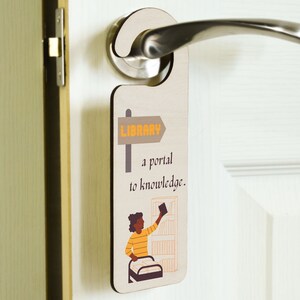 Può includere: Un appendiporta beige con una freccia marrone che punta a destra che dice "LIBRARY" in lettere arancioni. L'appendiporta ha anche un'illustrazione di cartone animato di una persona che spinge un carrello della biblioteca con libri e un cartello che dice "un portale alla conoscenza."