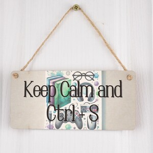 May include: A wooden sign with a white background and a colorful illustration of books, glasses, headphones, and a video game controller. The sign reads "Keep Calm and Ctrl+S".