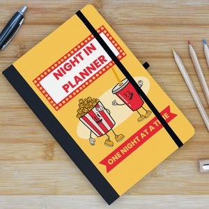 May include: A yellow notebook with a black elastic band closure. The cover features a cartoon illustration of popcorn and a soda cup with the text "Night in Planner" and "One Night at a Time".