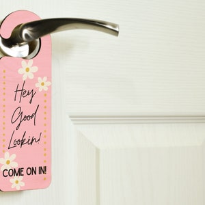 Könnte beinhalten: Rosafarbener Türanhänger mit dem Text "Hey Good Lookin'! COME ON IN!" in schwarzer Schrift. Der Türanhänger hat ein Blumendesign mit weißen Blumen und goldenen Punkten. Er hängt an einer weißen Tür.