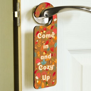 May include: A brown wooden door hanger with a fall-themed design. It features a variety of autumn leaves, mushrooms, and acorns in shades of red, orange, yellow, and brown. The text "Come in and Cozy Up" is printed in white on the hanger.