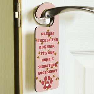 May include: Pink door hanger with gold glitter stars and the text "Please excuse the dog hair, it's our home's signature accessory". A red paw print with a white heart is at the bottom.