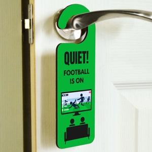 May include: Green door hanger with the text "Quiet! Football is on". The hanger features a cartoon image of a television with two people sitting on a couch watching a football game.