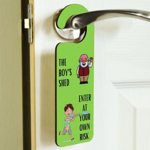 以下が含まれることがあります： 「The Boy's Shed」と「Enter at your own risk.」の文字が書かれた緑色のドアハンガー。ハンガーには、ひげを生やした男性と水鉄砲を持った少年の漫画が描かれている。