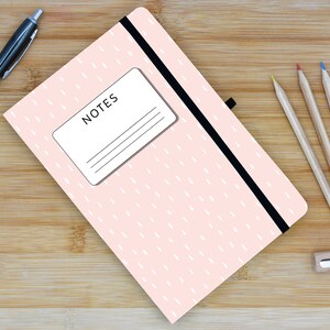 May include: A pink notebook with a black elastic band closure and a white label that says "NOTES". The notebook is on a wooden surface with a pen and pencils.