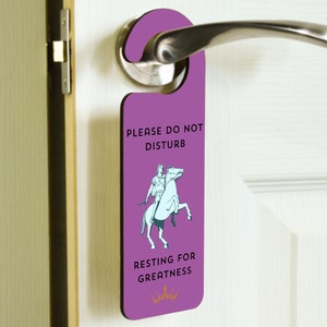 Op de afbeelding: Paarse deurhanger met een witte illustratie van een persoon die op een paard rijdt. De tekst luidt "Alstublieft niet storen, rusten voor grootheid".