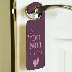 Op de afbeelding: Paarse deurhanger met de tekst "Niet storen" in roze letters. De hanger heeft ook een roze voetafdrukdesign en kleine roze hartjes.