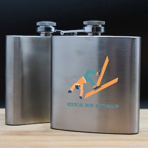 May include: Two silver hip flasks with a hinged lid. One flask has a graphic of a person skiing upside down with the text "Vertical Drop, Bottom Up".
