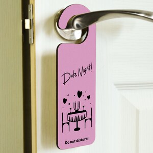 Op de afbeelding: Roze deurhanger met de tekst "Date Night!" en een zwart silhouet van een tafeldekking met kaarsen en harten. De tekst "Niet storen!" staat onderaan.