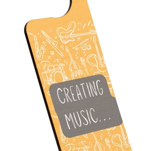 Pode incluir: Um porta-chaves de madeira amarelo com um contorno branco de instrumentos musicais e o texto "Creating Music..." em um quadro-negro cinza.