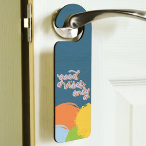 Peut inclure: Un porte-manteau de porte bleu avec le texte "good vibes only" dans une police d'écriture manuscrite colorée. Le porte-manteau a un design abstrait coloré avec des éclaboussures de peinture orange, jaune, verte et bleue.