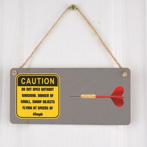 Puede incluir: Un cartel de precaución amarillo y negro con el texto "CAUTION DO NOT OPEN WITHOUT KNOCKING. DANGER OF SMALL, SHARP OBJECTS FLYING AT SPEEDS OF 40mph" colgado en una puerta de madera gris con un dardo rojo y dorado.