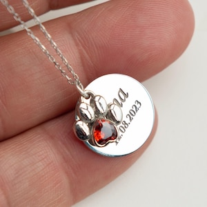 Può includere: Collana con pendente in argento con un'impronta di zampa con una gemma rossa e un ciondolo circolare inciso. La collana ha una catena delicata.