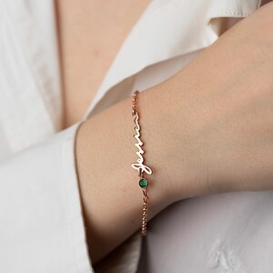 May include: A rose gold chain bracelet with a cursive name charm and a small green gemstone.