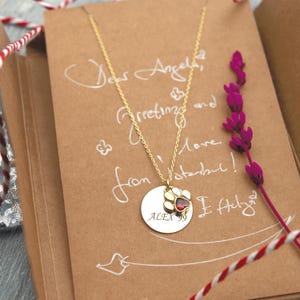 May include: A gold necklace with a circular pendant displaying a paw print with a red gemstone. The pendant is engraved with "ALEX 99". The necklace rests on a brown card with handwritten text and a sprig of purple flowers.