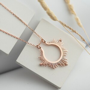 May include: A rose gold necklace featuring a sunburst pendant. The pendant is a circle with rays of light radiating from it. The necklace is displayed on a white surface.