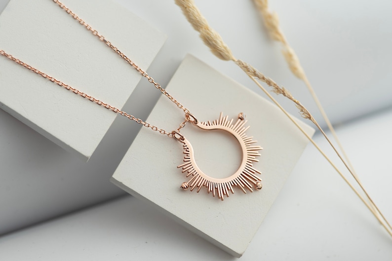 May include: A rose gold necklace featuring a sunburst pendant. The pendant is a circle with rays of light radiating from it. The necklace is displayed on a white surface.