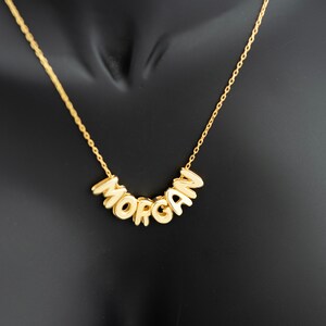 Könnte beinhalten: Eine goldfarbene Halskette mit dem Namen "MORGAN" in Blockbuchstaben. Die Buchstaben sind verbunden und leicht gebogen. Die Halskette wird vor einem schwarzen Hintergrund gezeigt.