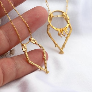 Könnte beinhalten: Zwei goldfarbene Halsketten mit einzigartigen, von der Natur inspirierten Designs. Eine zeigt eine stilisierte Herzform, die andere ein ringförmiges Design mit einem zentralen Edelstein. Beide sind an filigranen Ketten.